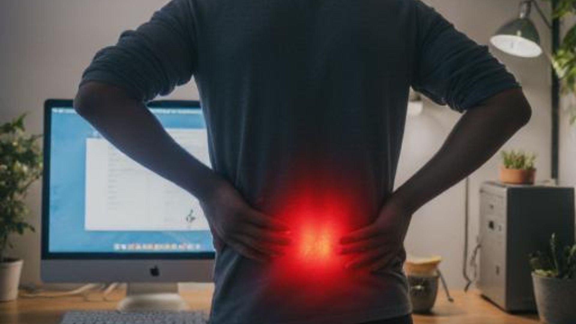 A man at a desk presses his hand into his lower back in an office setting, showing strain after a long day